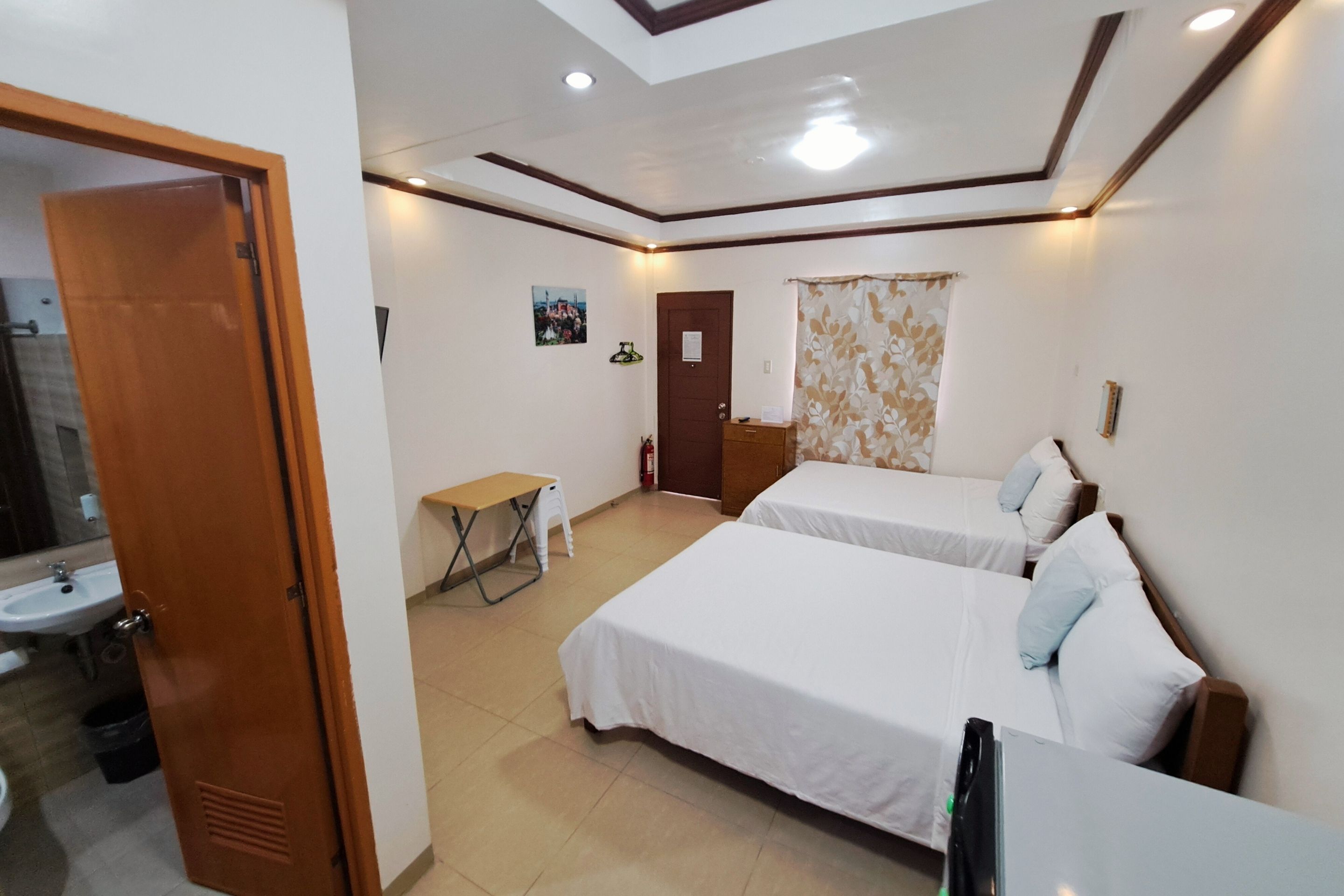 A hotel room with two beds, a desk, a chair, a small table, a bathroom with a sink, and a door leading to another room.