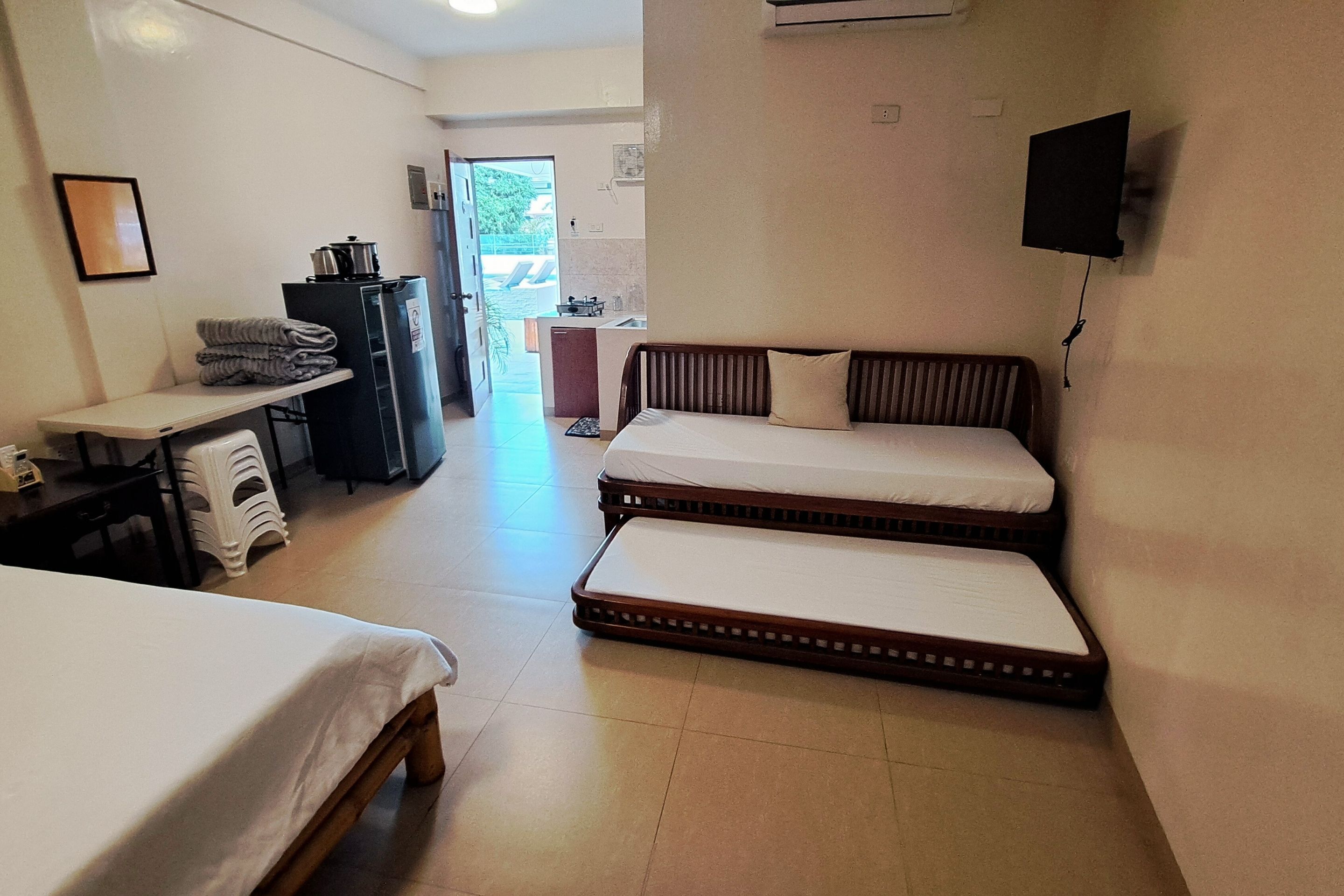 Une chambre d’hôtel avec deux lits simples, un bureau avec une chaise, une petite kitchenette et un téléviseur fixé au mur.