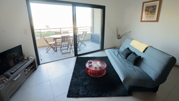 Un salon comprenant un canapé, une table basse, un téléviseur et une porte-fenêtre coulissante donnant sur un balcon équipé d’une table et de chaises.