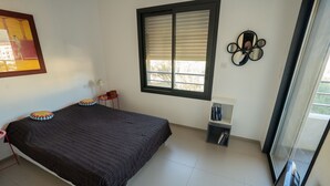 Une chambre à coucher avec un lit, une fenêtre avec des stores, une petite table avec une chaise et une étagère fixée au mur, sur laquelle sont posés divers objets.