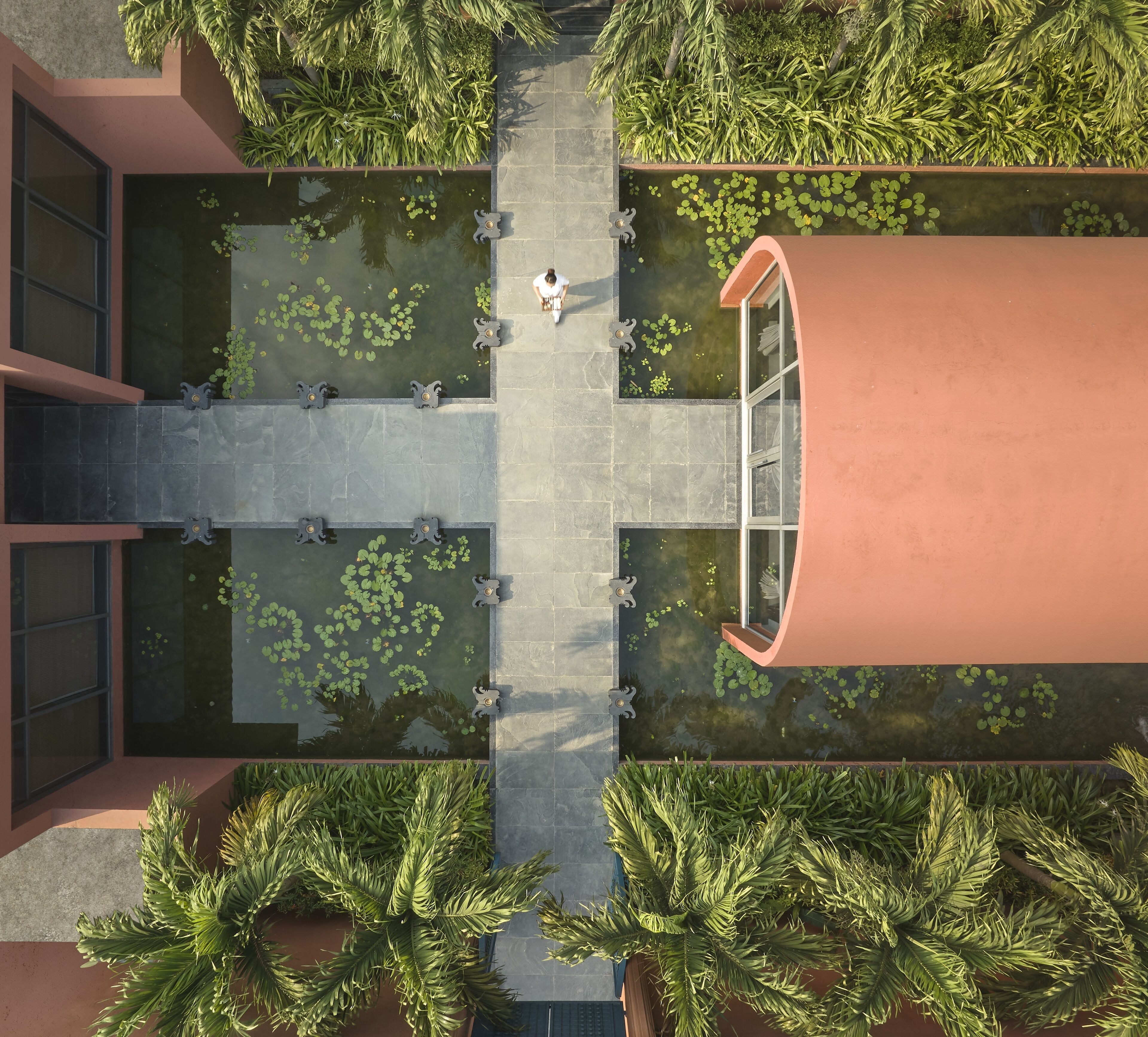 An aerial view of a modern building with a central courtyard featuring a water garden, surrounded by lush greenery and a curved structure.
