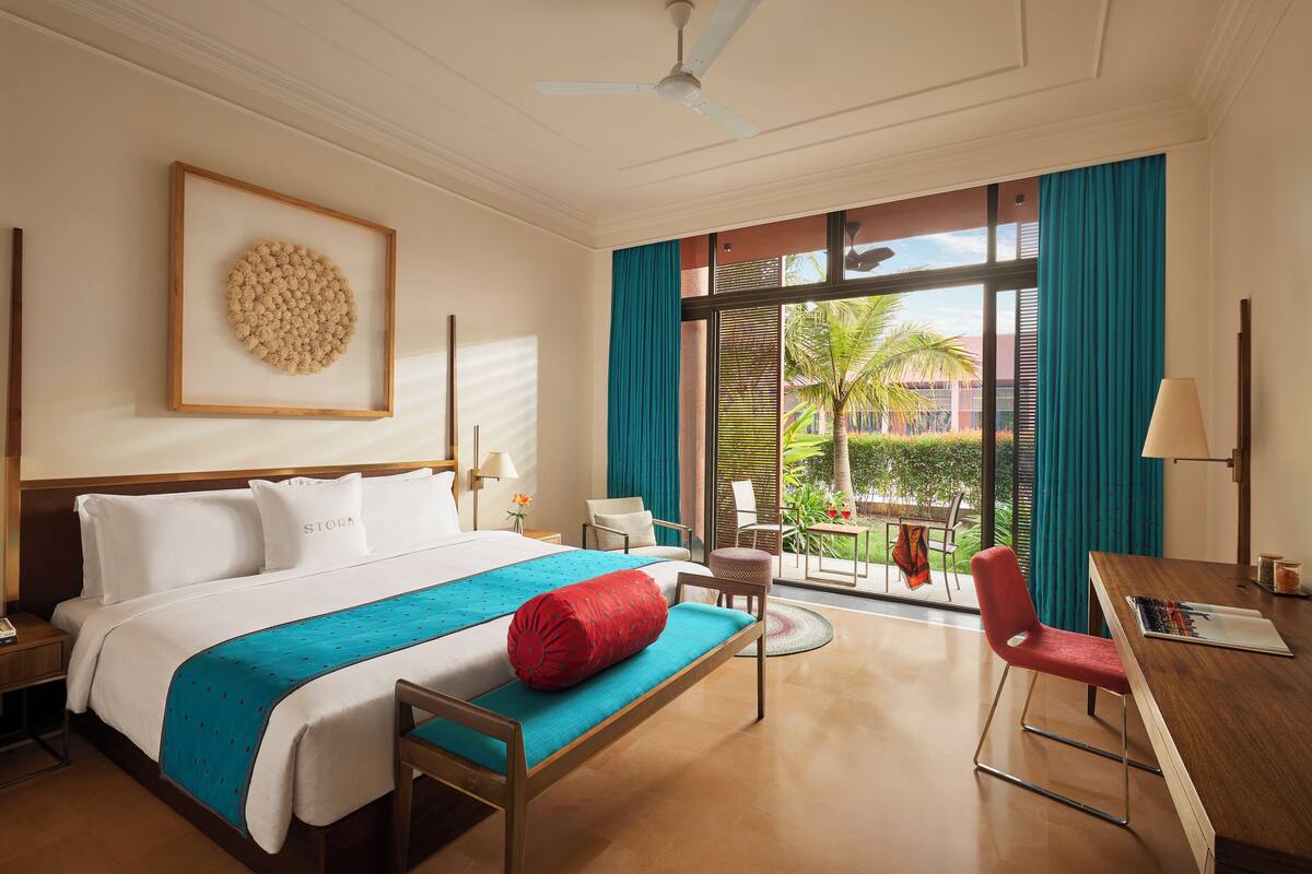 Quarto de hotel com cama, escrivaninha, cadeira, janela com cortinas e ventilador de teto.