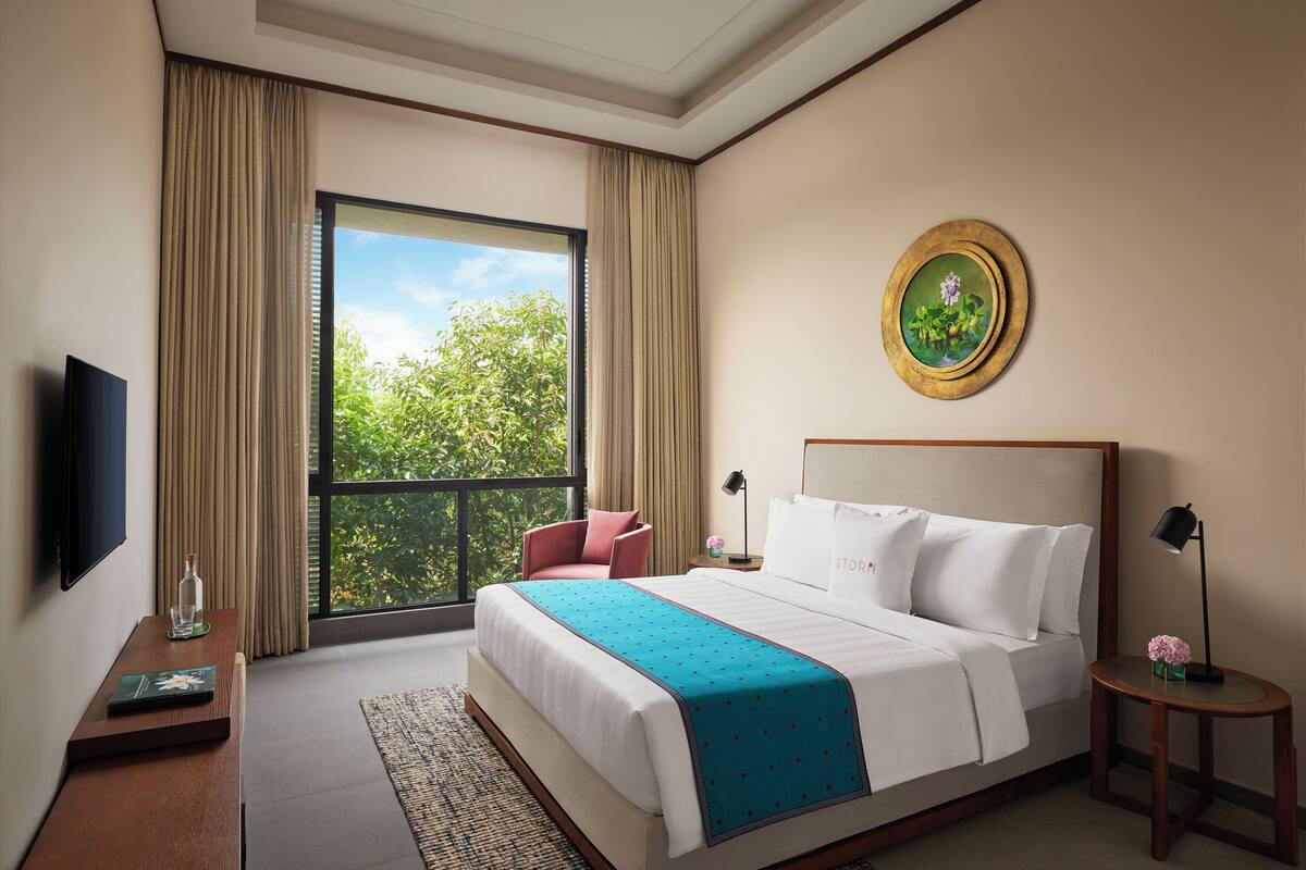 Quarto de hotel com uma cama grande, televisão, janela com cortinas, um quadro redondo e uma pequena mesa com abajur.