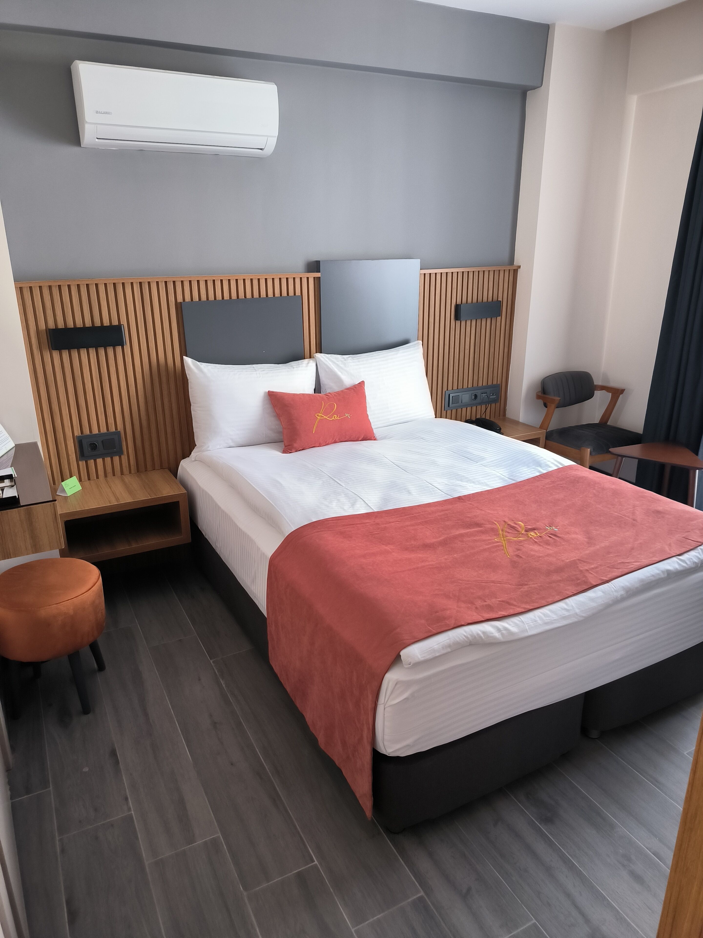 A modern hotel room with a large bed, a wooden headboard, a bedside table, a chair, and an air conditioning unit.