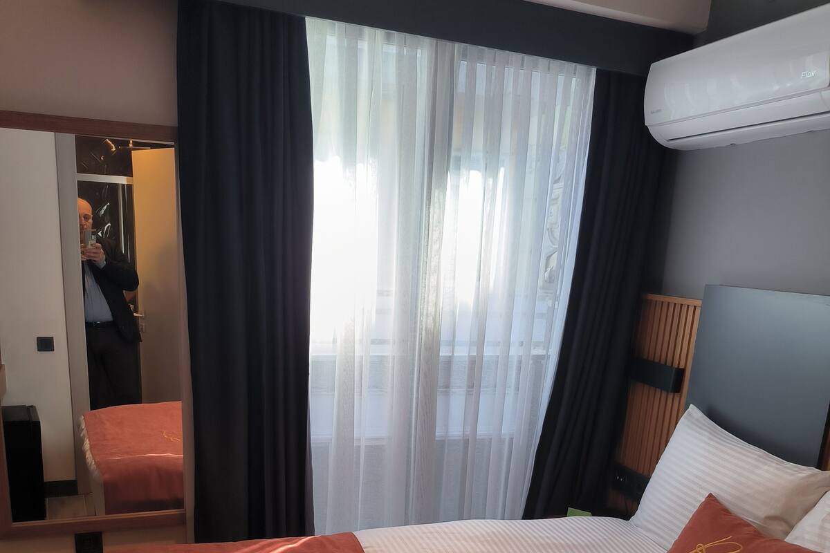 Habitación de hotel con una cama grande, un espejo, una ventana con cortinas y un aire acondicionado instalado en el techo.