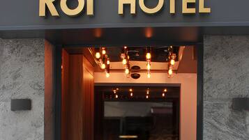 The entrance of Roi Hotel with a modern design, featuring a wooden door, a black mat with the hotel name, and two potted plants on either side.