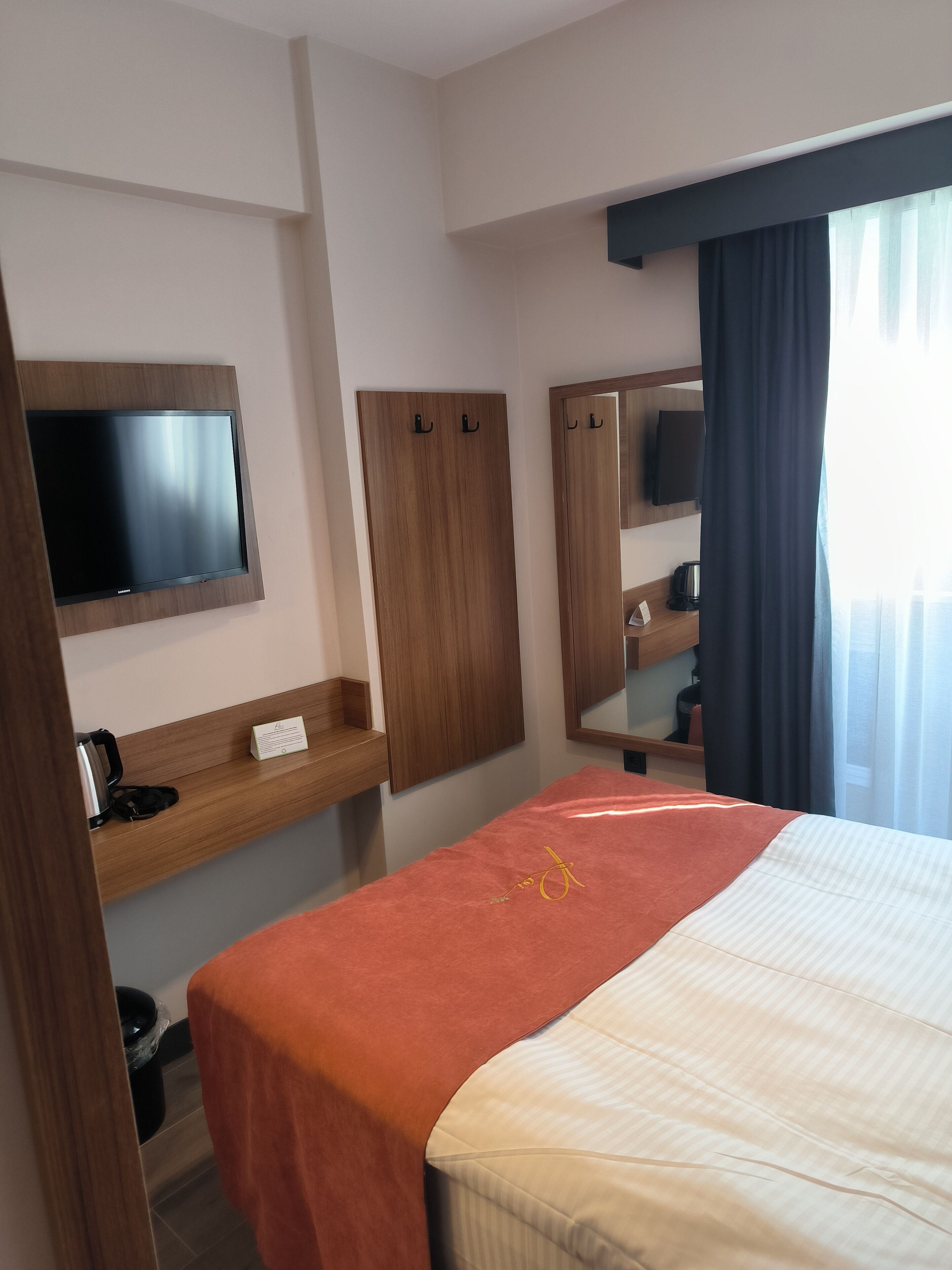 A hotel room with a bed, a TV mounted on the wall, a wooden wardrobe, a mirror, and a window with curtains.