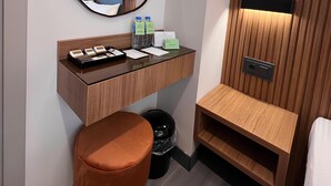 A modern hotel room with a wooden vanity, a round mirror, a small stool, a trash bin, and a wooden cabinet with a built-in heater. - Roi Hotel Izmir (Izmir)