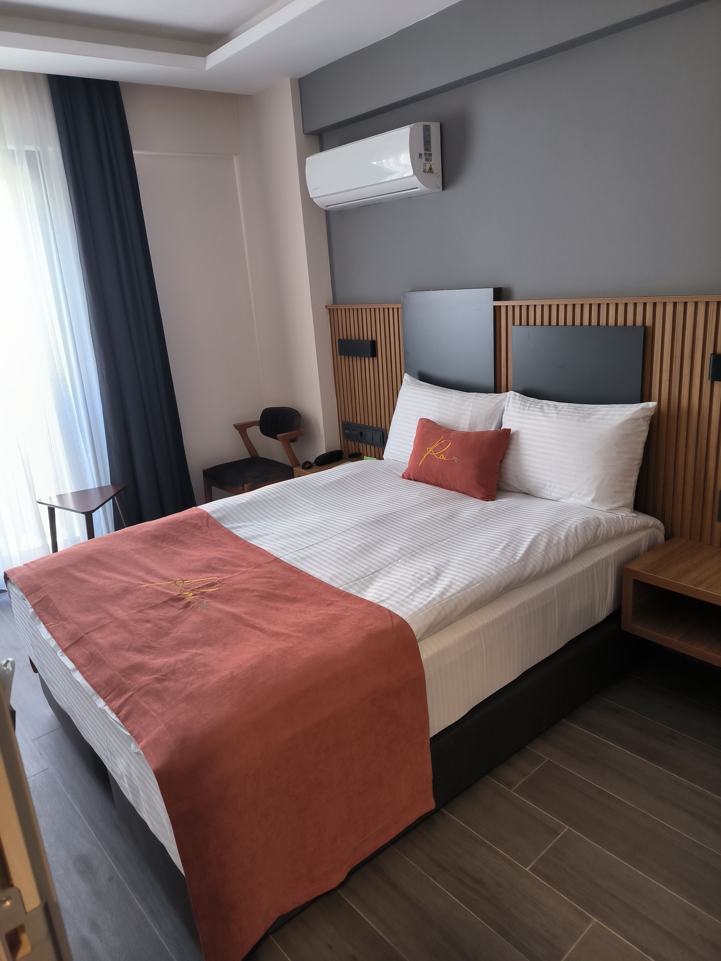 A hotel room with a bed, a nightstand, a chair, an air conditioning unit, and a window with curtains.