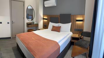 A hotel room with a large bed, a bedside table, a chair, a small table, a mirror, and an air conditioning unit.