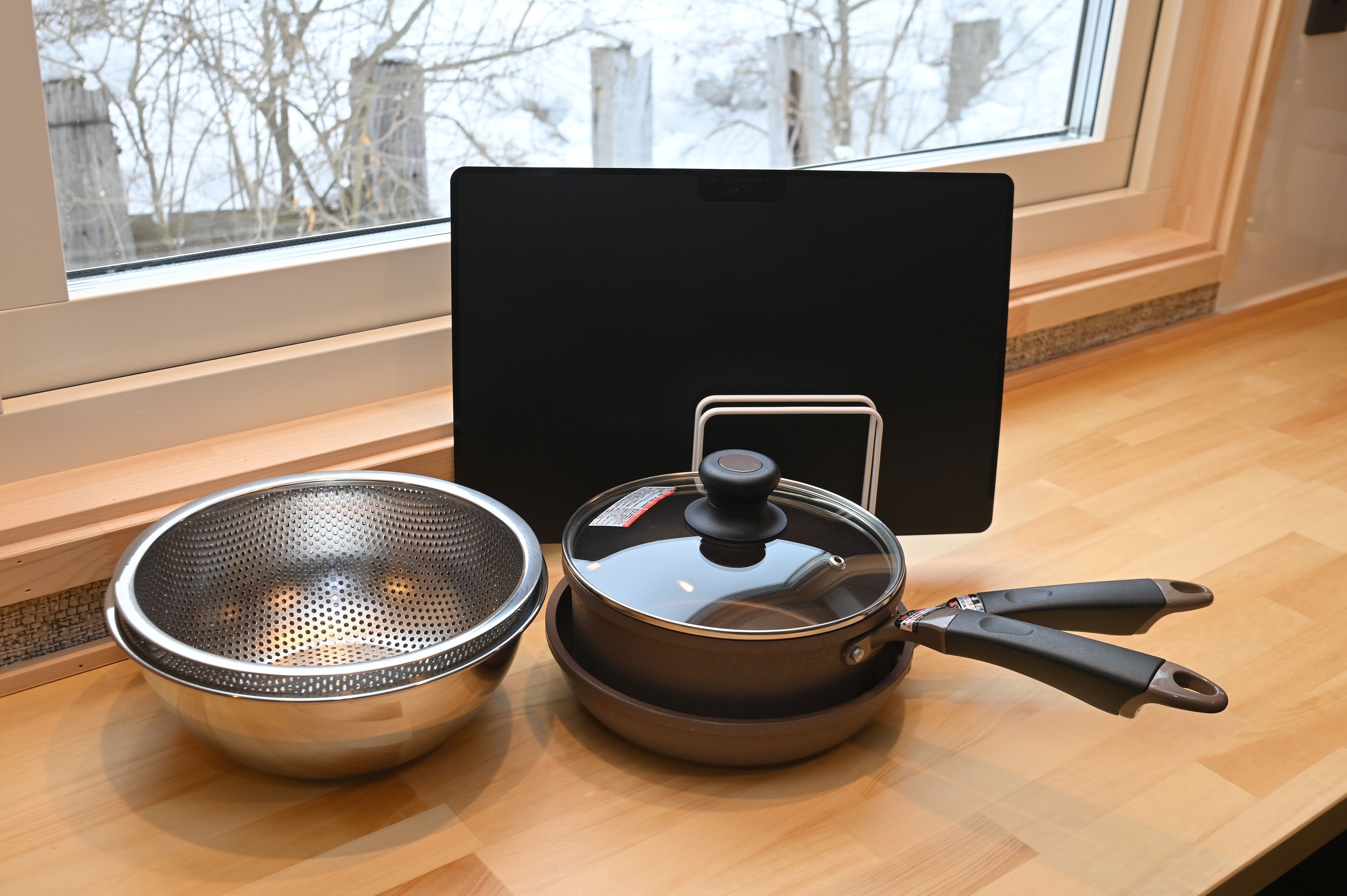A kitchen with a window, a colander, a pot with a lid, and a pair of tongs on a wooden countertop.