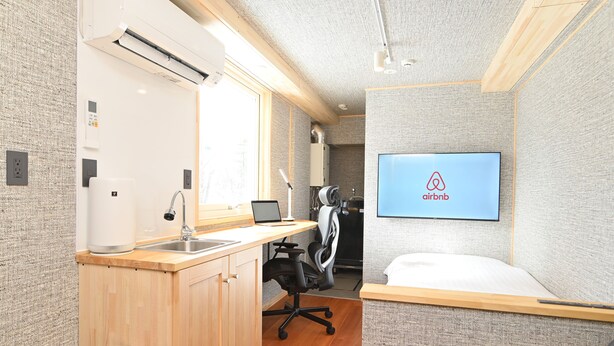Habitación compacta con kitchenette, una cama, un escritorio y un televisor que muestra el logo de Airbnb.
