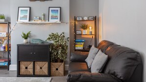 Una sala de estar moderna con un sofá de cuero negro, un mueble de madera con cestas tejidas, una planta en maceta y una estantería con marcos de fotos y adornos.