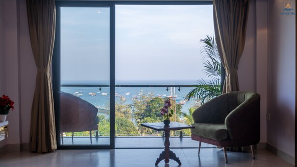A room with a large sliding glass door leading to a balcony with a view of the sea and boats. There are two chairs and a small table in the room.