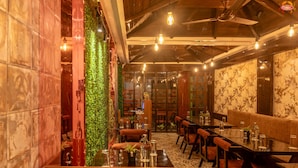 A restaurant interior with wooden beams, hanging light bulbs, and a green wall. Tables and chairs are arranged for dining.
