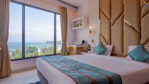 A hotel room with a large bed, a desk with a coffee maker, a shelf with decorative items, and a view of the ocean.