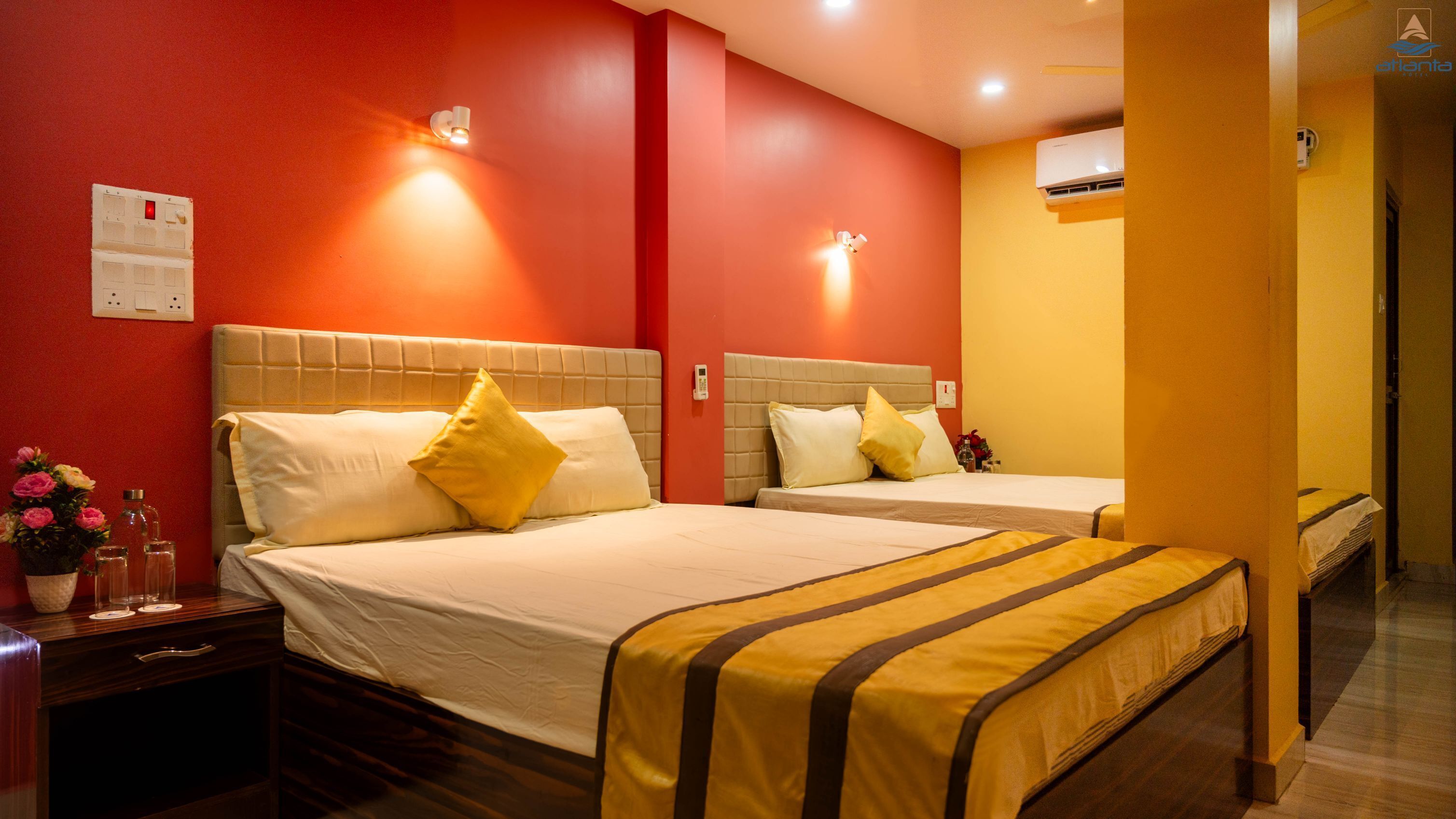 A hotel room with two beds, a nightstand with a vase of flowers, and a wall-mounted air conditioning unit.