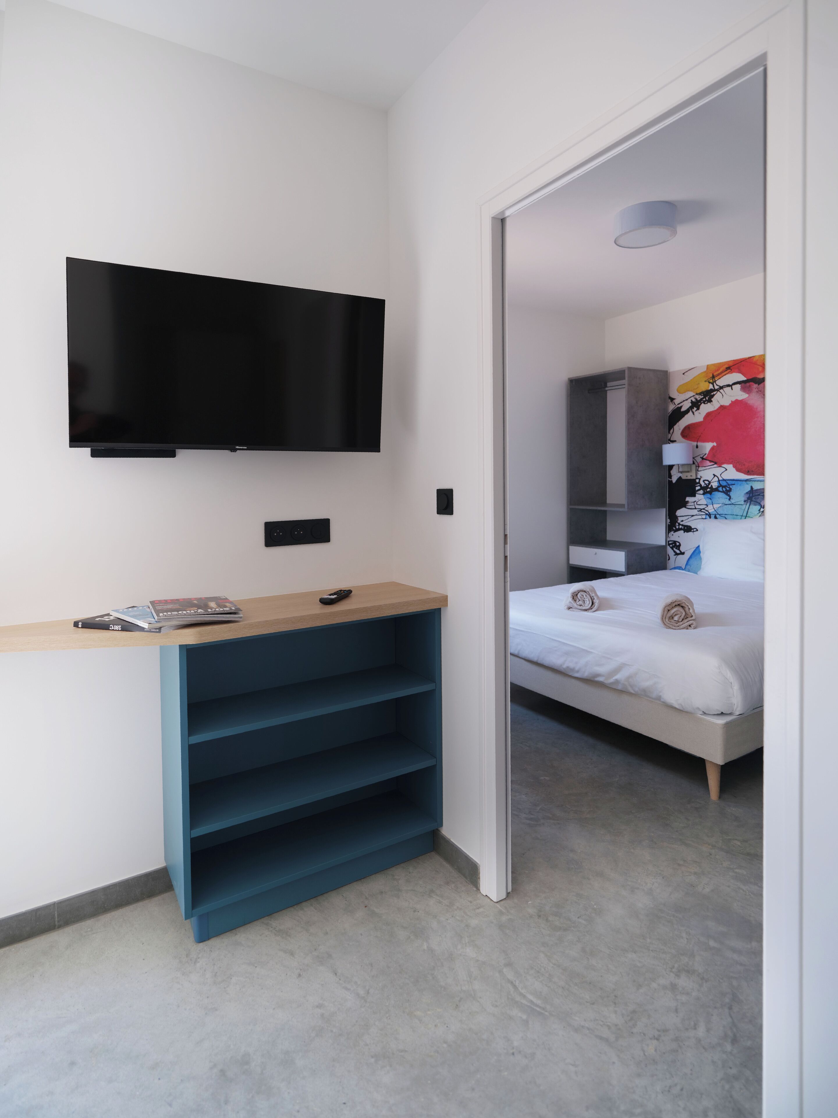Une chambre moderne dotée d’un téléviseur à écran plat fixé au mur, d’un bureau en bois avec une télécommande et d’un lit agrémenté de serviettes soigneusement pliées.