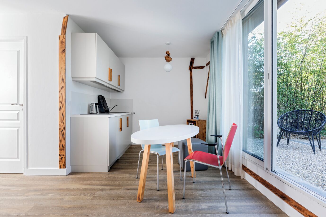 A compact kitchen with white cabinets, a dining table with two chairs, and a large window with a view of greenery.