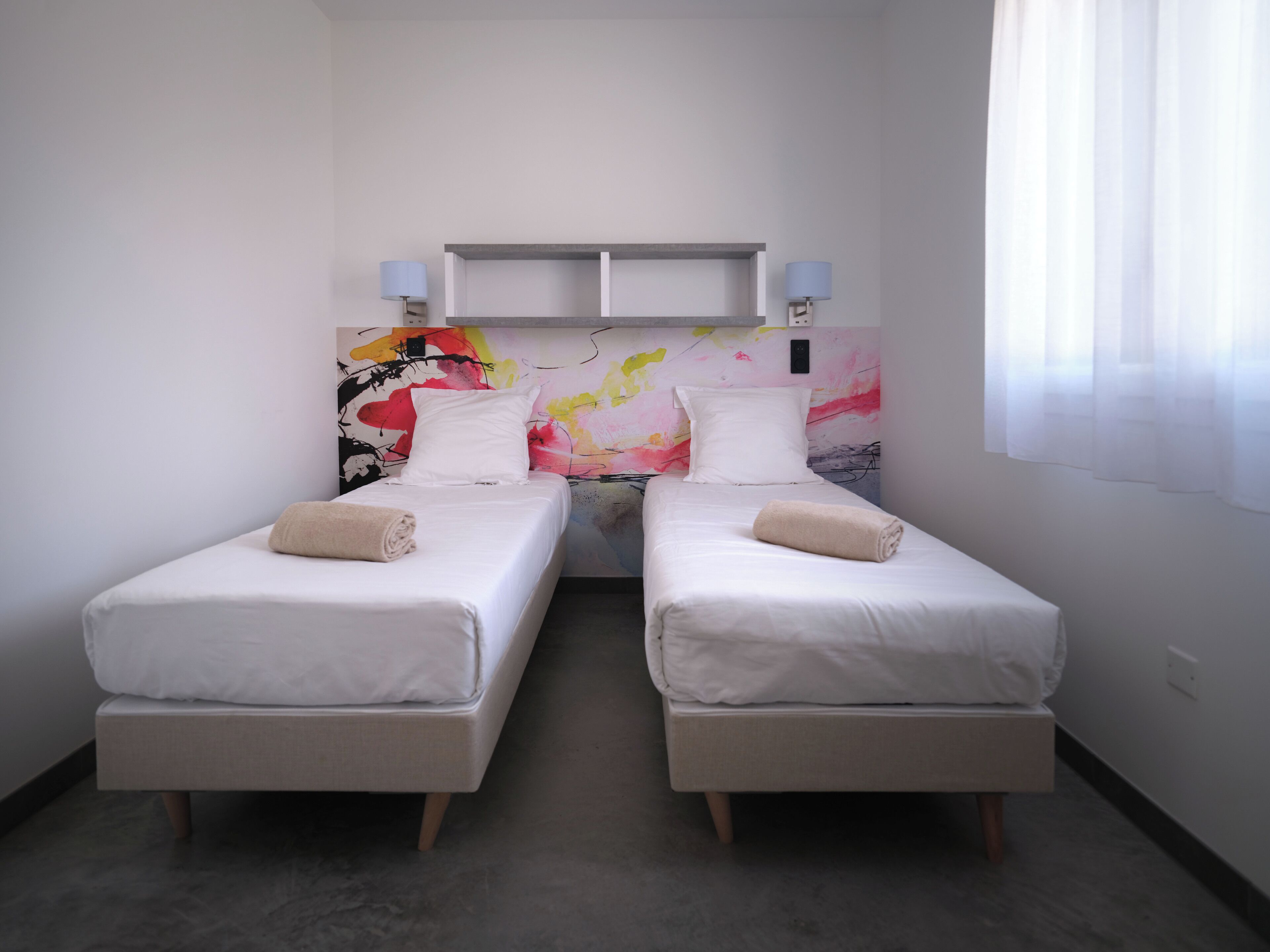 Une chambre d’hôtel avec deux lits simples, une peinture abstraite aux couleurs vives accrochée au mur et une fenêtre ornée de rideaux légers.