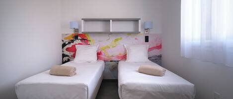 Quarto de hotel com duas camas de solteiro, uma pintura abstrata colorida na parede e uma janela com cortinas leves.
