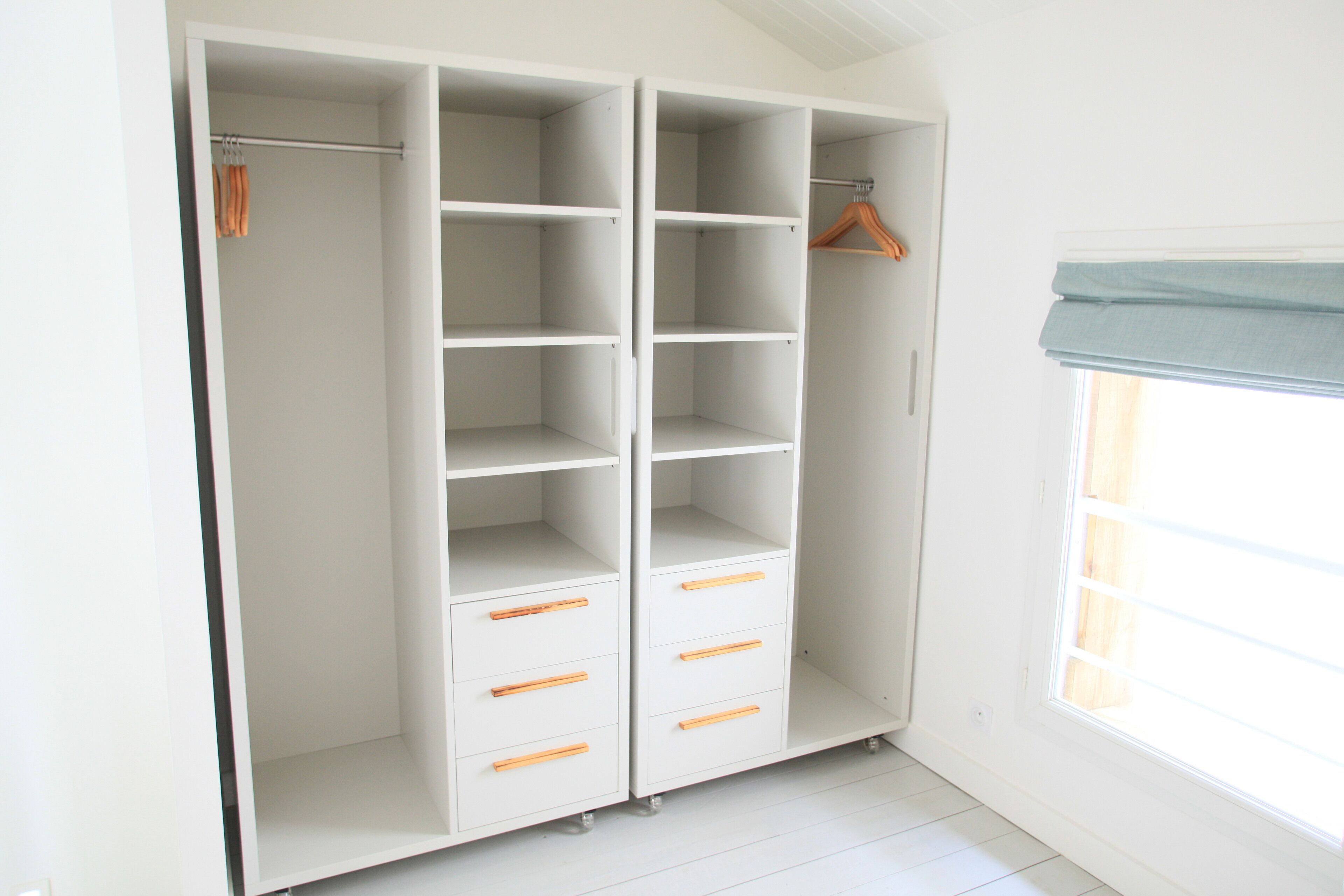 Un closet amplio y blanco con varias estanterías y cajones, un perchero de madera y una ventana con una persiana azul.