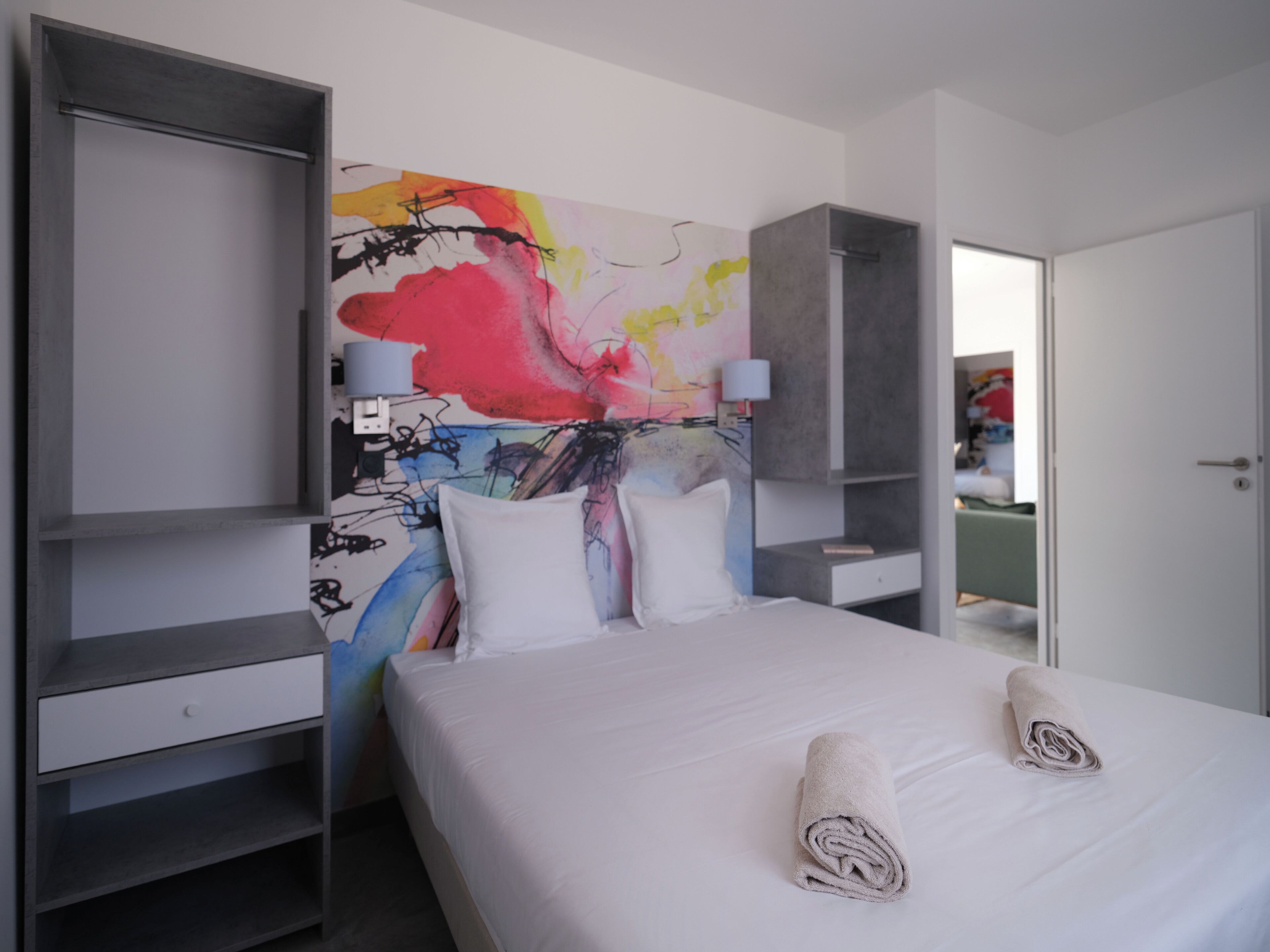 A modern hotel room with a large bed, white bedding, and a colorful abstract painting on the wall.
