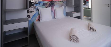A modern hotel room with a large bed, white bedding, and a colorful abstract painting on the wall.