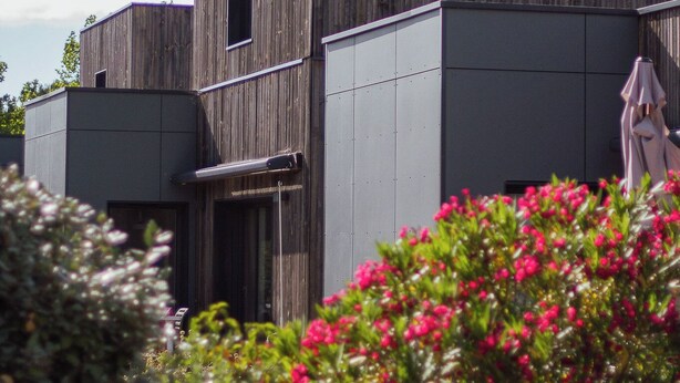 A modern building with a wooden facade and a grey section, surrounded by greenery and flowers. - Côté Océan (Angoulins)