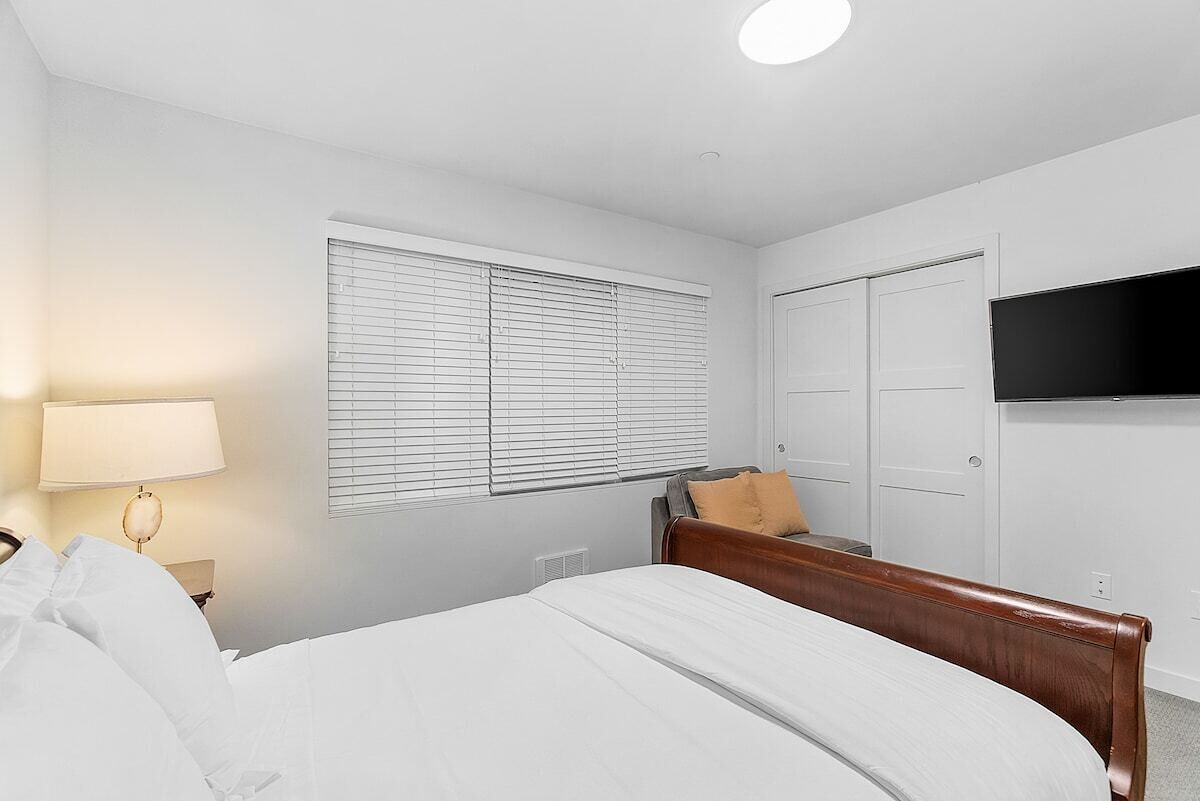 A hotel room with a bed, a TV mounted on the wall, a window with blinds, and a door.