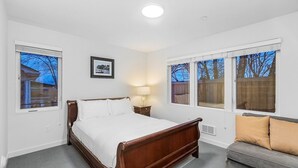 A bedroom with a wooden bed, a gray sofa, and large windows with a view of trees and a house. - The Sweetest Hideaway - 81 Walkscore! (Seattle)