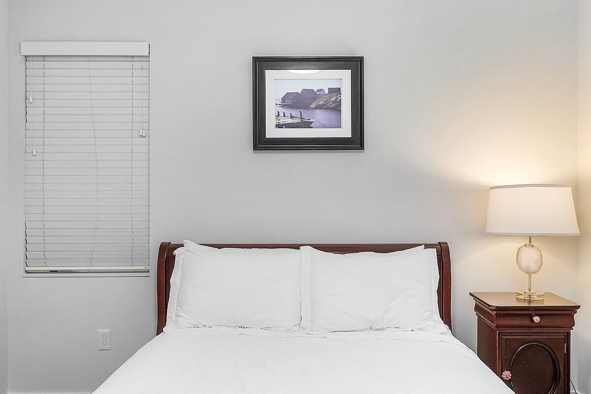 Un letto ben rifatto con lenzuola bianche, un quadro appeso al muro, una lampada da comodino e una finestra con persiane.