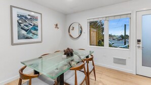 A dining area with a glass table, wooden chairs, a framed artwork, a round mirror, and a window with a view of a parking lot and trees. - The Sweetest Hideaway - 81 Walkscore! (Seattle)