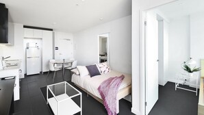 Une chambre d’hôtel moderne et minimaliste, comprenant un lit, un petit bureau, une chaise, une table basse en verre et un téléviseur.