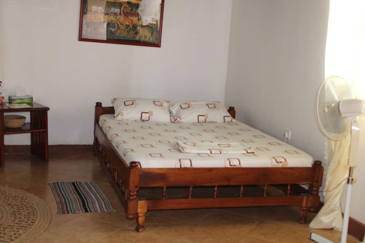 A bedroom with a wooden bed, a bedside table, a wall-mounted fan, and a patterned rug.