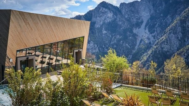 A modern building with large glass windows, surrounded by a pool and lounge chairs, set against a mountainous backdrop. - Pure Thermal Résidence (Roquebilliere)