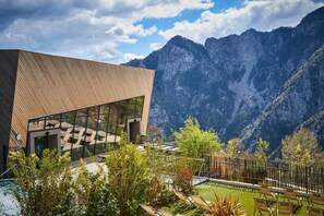 A modern building with large glass windows, surrounded by a pool and lounge chairs, set against a mountainous backdrop. - Pure Thermal Résidence (Roquebilliere)