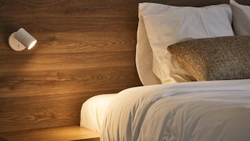 A neatly made bed with white linens and a textured pillow, a wooden nightstand with a wall-mounted light, and a wooden headboard.