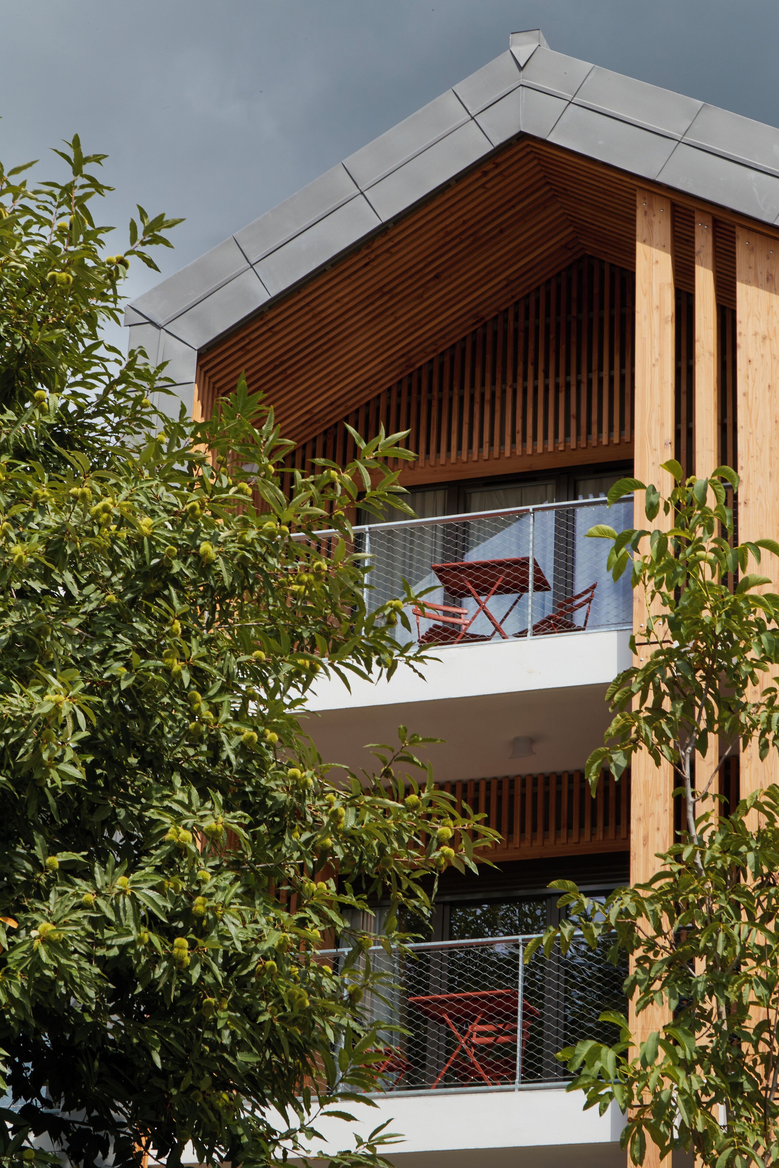 Een modern gebouw met een houten gevel, een balkon met rode meubels, en een boom op de voorgrond.