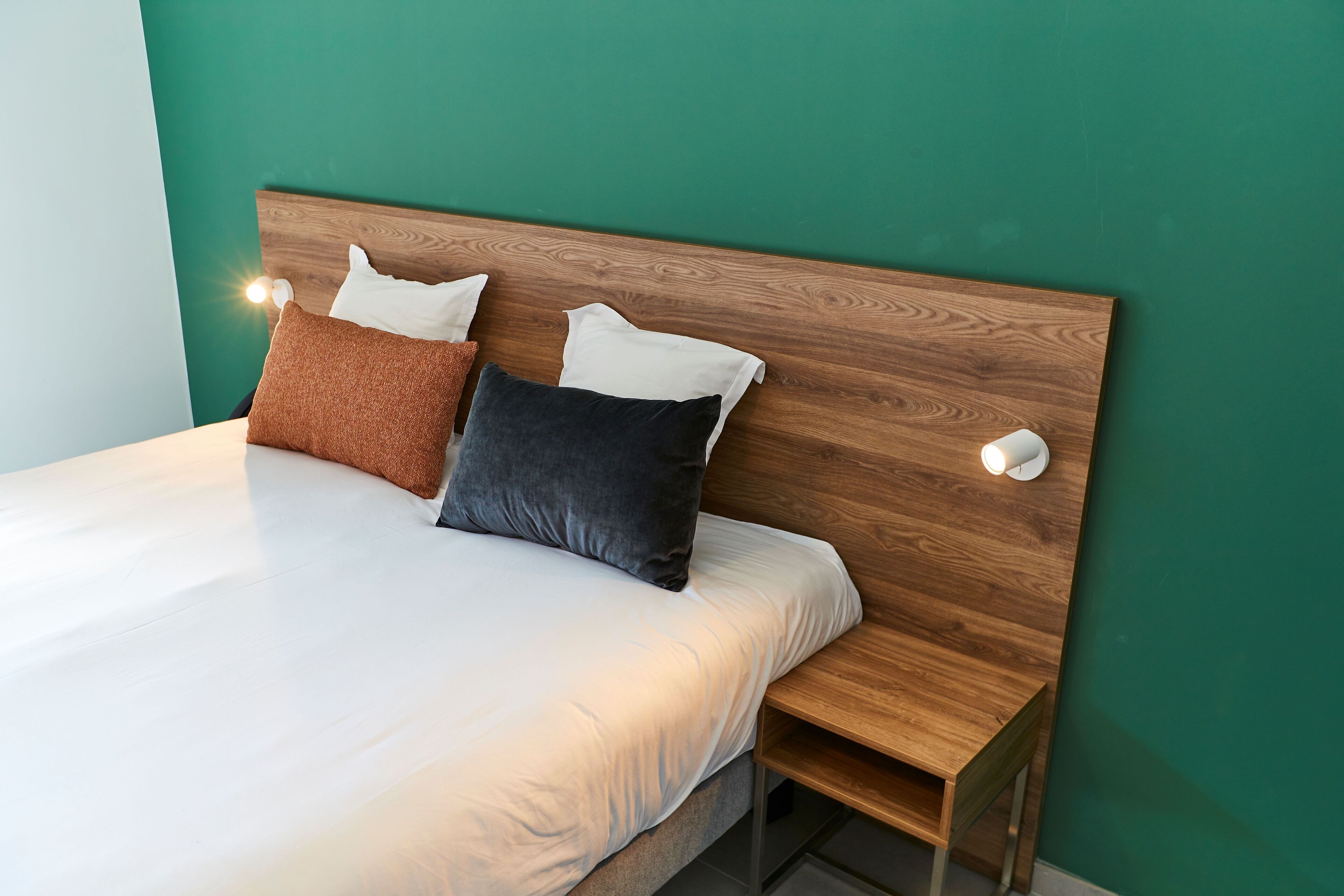 Een net opgemaakt bed met witte lakens en twee kussens, een houten hoofdbord en een nachtkastje met een lamp.