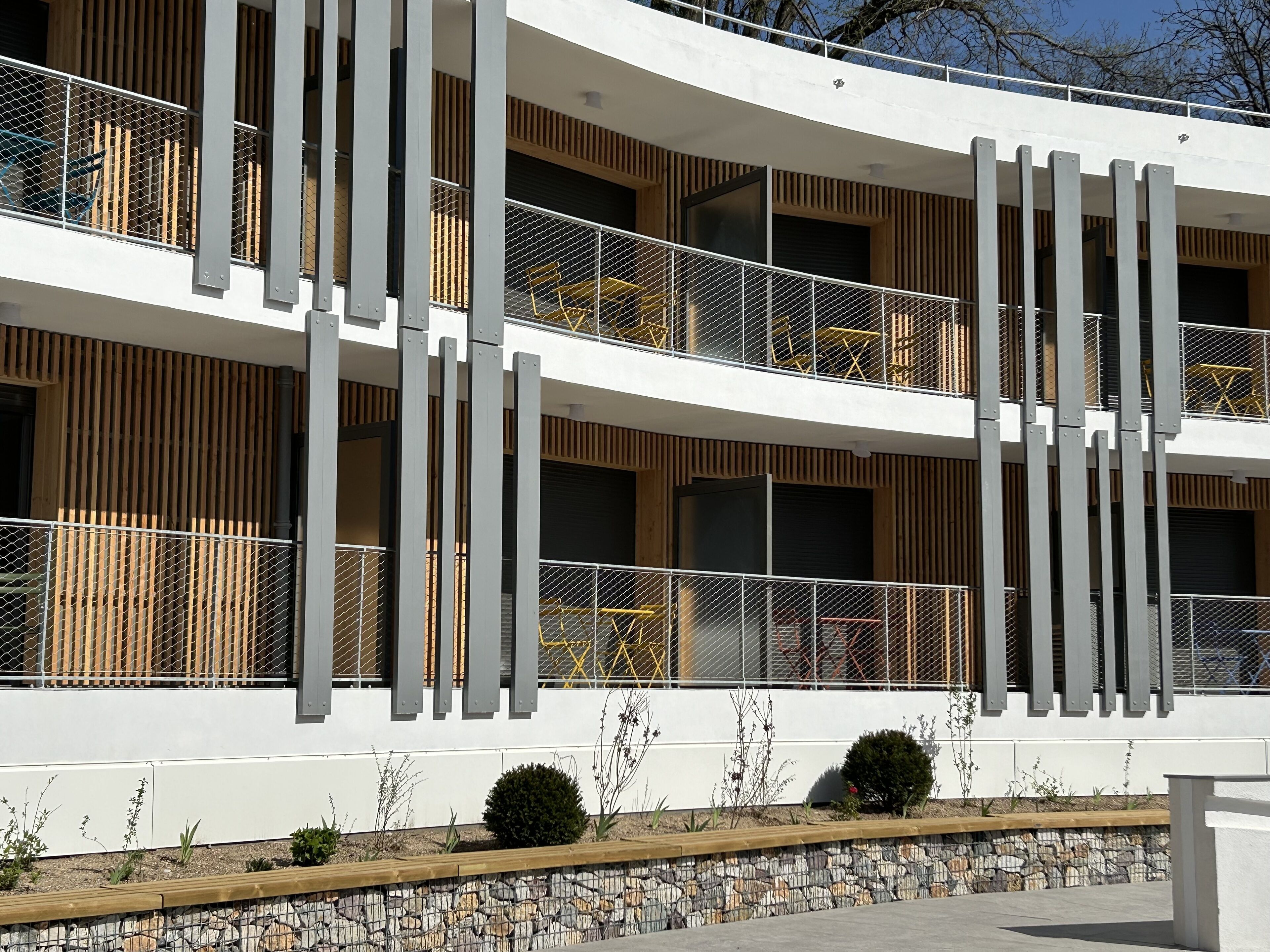 A modern multi-story building with balconies, a stone retaining wall, and minimalistic landscaping.