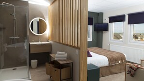 A modern hotel room with a glass-enclosed shower, a round mirror, a wooden partition, a bed with a view, and a small table with a chair.