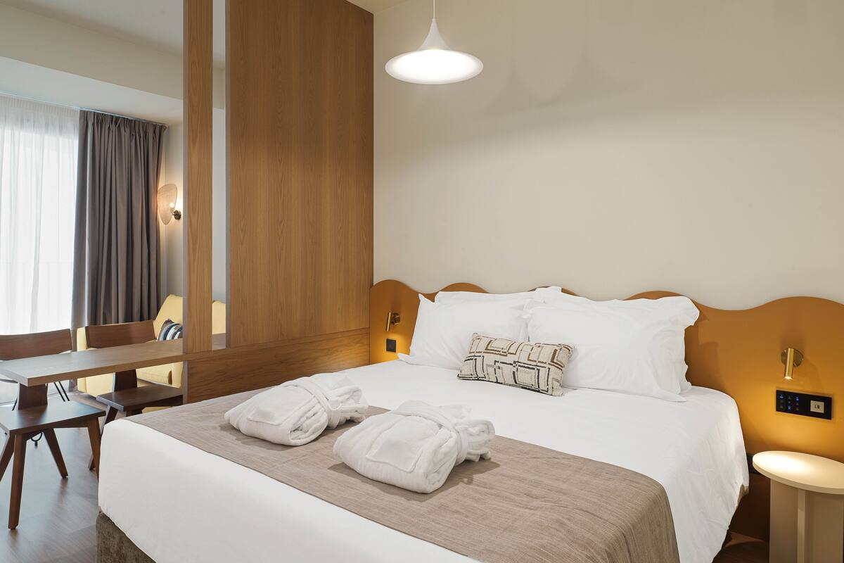 Quarto de hotel com cama arrumada, cabeceira de madeira, mesa de cabeceira, escrivaninha e um espelho grande.