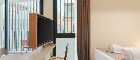A hotel room with a wooden desk, a television, a bed with white linens, a window with blinds, and a view of buildings outside.