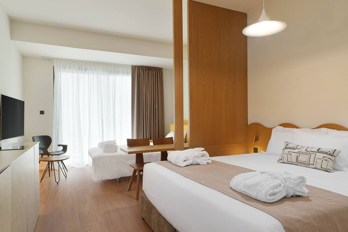 Quarto de hotel com cama, escrivaninha, cadeira, televisão e uma janela grande com cortinas.