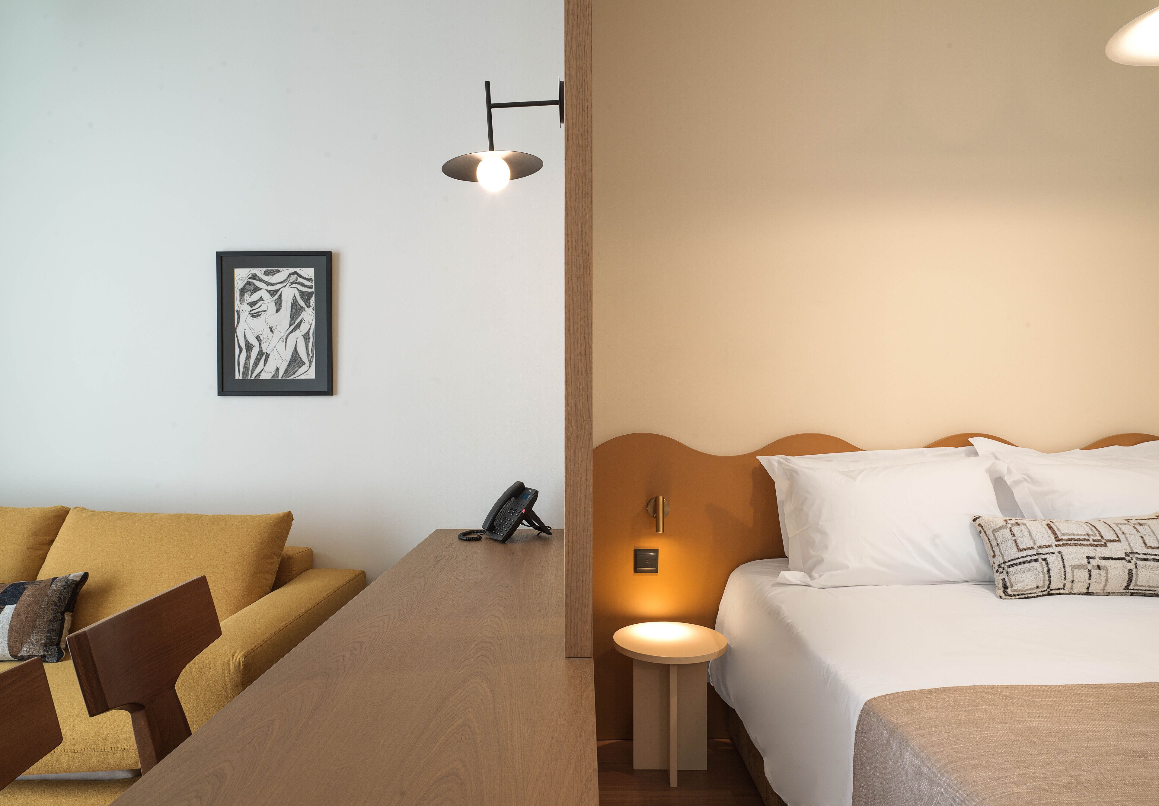 Habitación de hotel con un escritorio de madera, una cama con ropa blanca, una mesita de noche con lámpara y un cuadro en la pared.