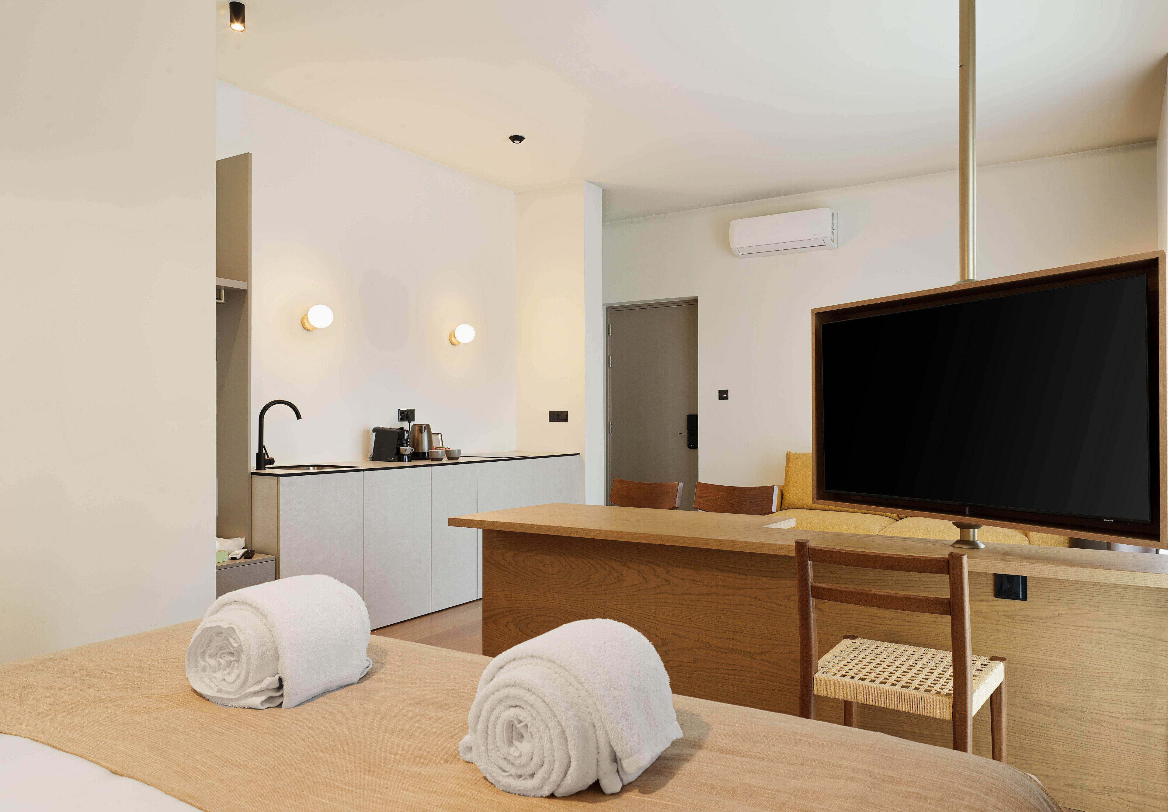 A hotel room with a bed, a desk, a chair, a TV, a kitchen area, and a bathroom.