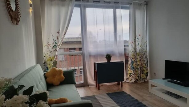 A living room with a sofa, a TV, a small table, and a view through a window. - Appartement aux Portes de Paris, Métro 12 (Aubervilliers)