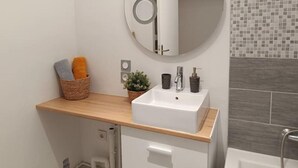 A modern bathroom with a round mirror, white sink, and a wooden shelf with towels and a plant. - Appartement aux Portes de Paris, Métro 12 (Aubervilliers)