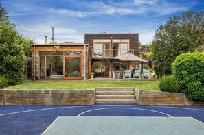 A modern two-story house with a patio area, a basketball court, and a wooden deck.